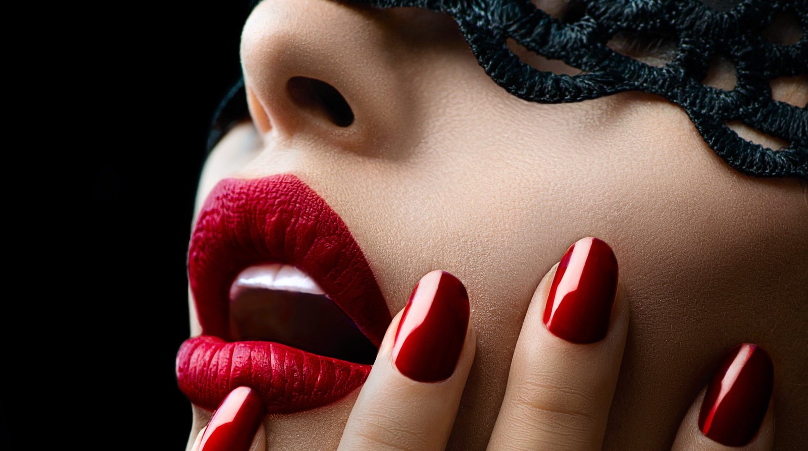 Woman's face, masked in black lace, red lipstick, fingers up on her cheek, fingers tipped in red polished nails.